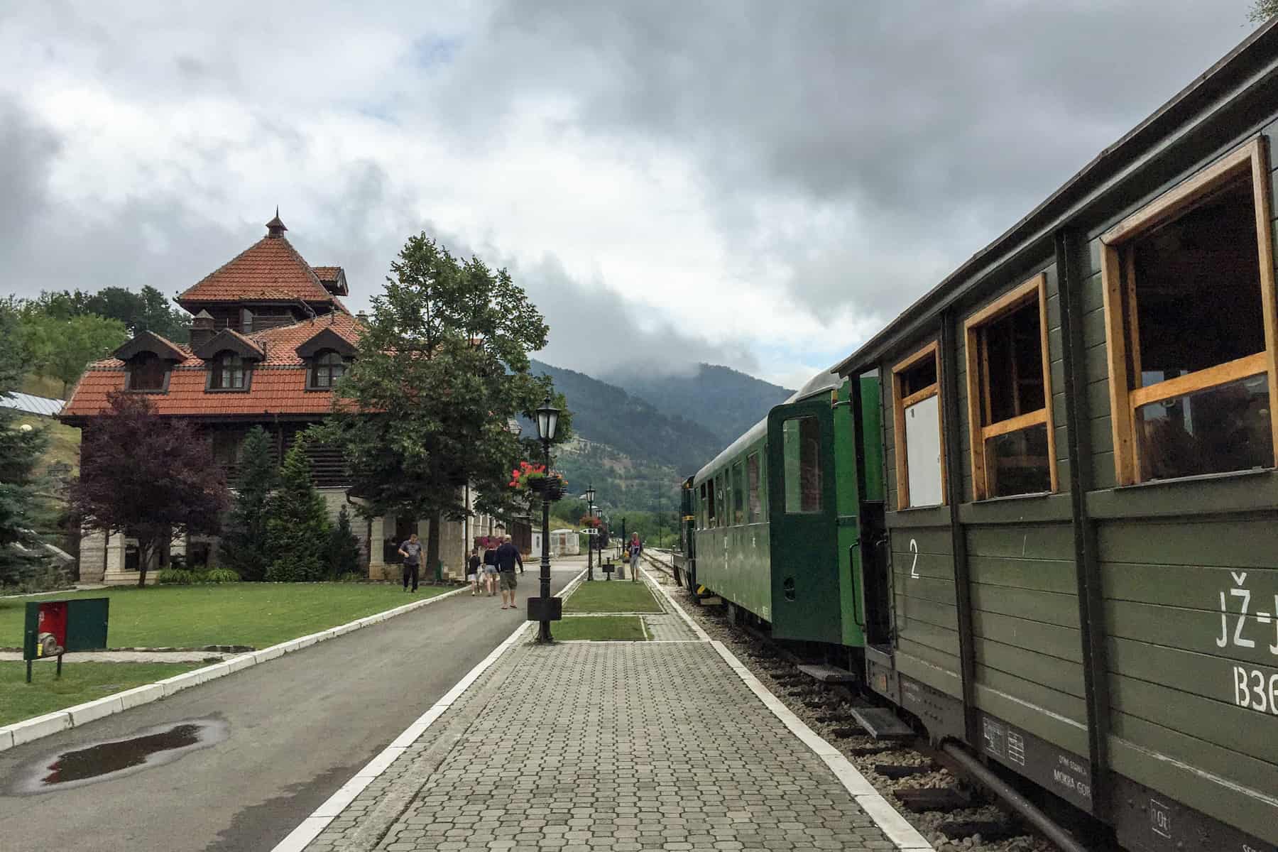 A dark green wood panelled train - The Sargan Eight Railway in Serbia - at the station platform with orange roofed buildings, in the direction of the mountains.