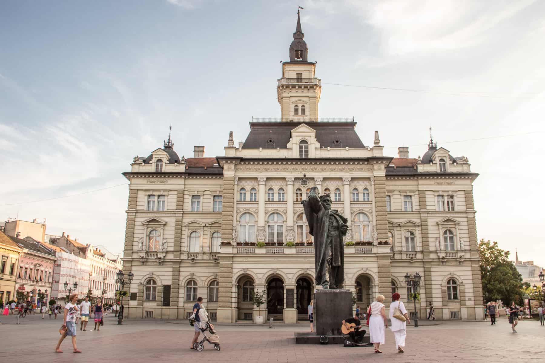 A dark, tall statue of a man in a public square and in front of a classical, cream coloured building with a silver roof spire.. 