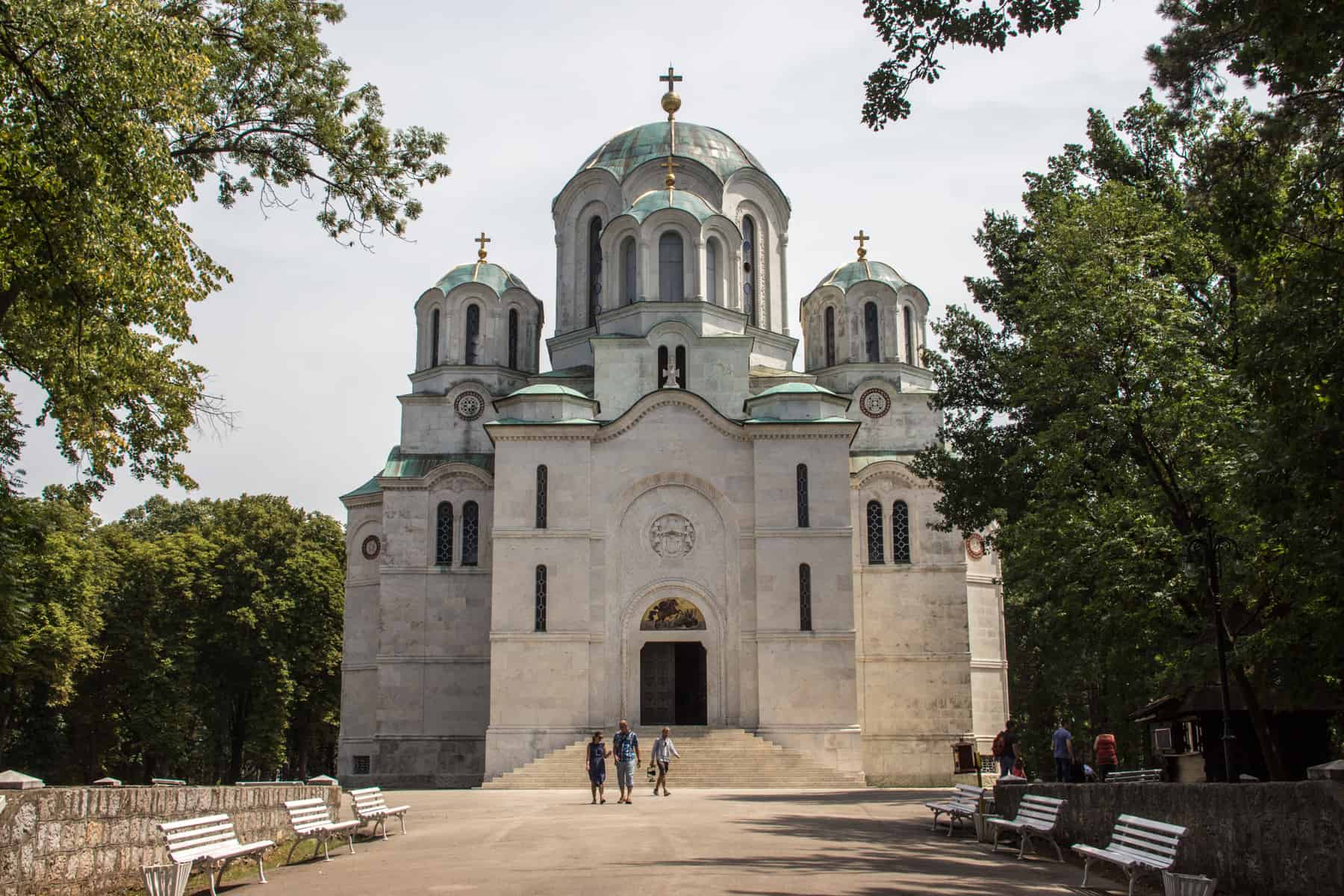 Three people walk outside a large white marble church, with six mint green domes, four of which are topped with gold crosses.