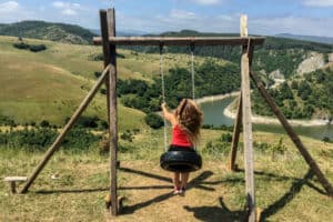 A woman with long hair, wearing a red top, sits of a tyre swing overlooking a winding gorge surrounded by hills and forest.