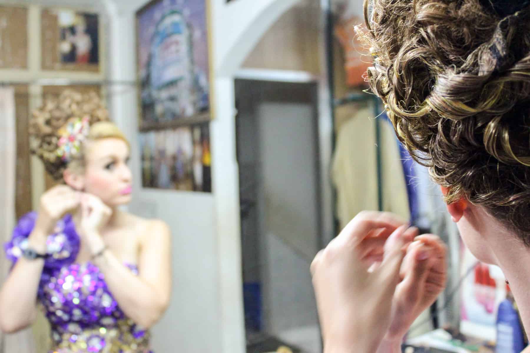 A blonde woman in a purple dress looks into a mirror and applies an earring during a photoshoot in Cambodia.