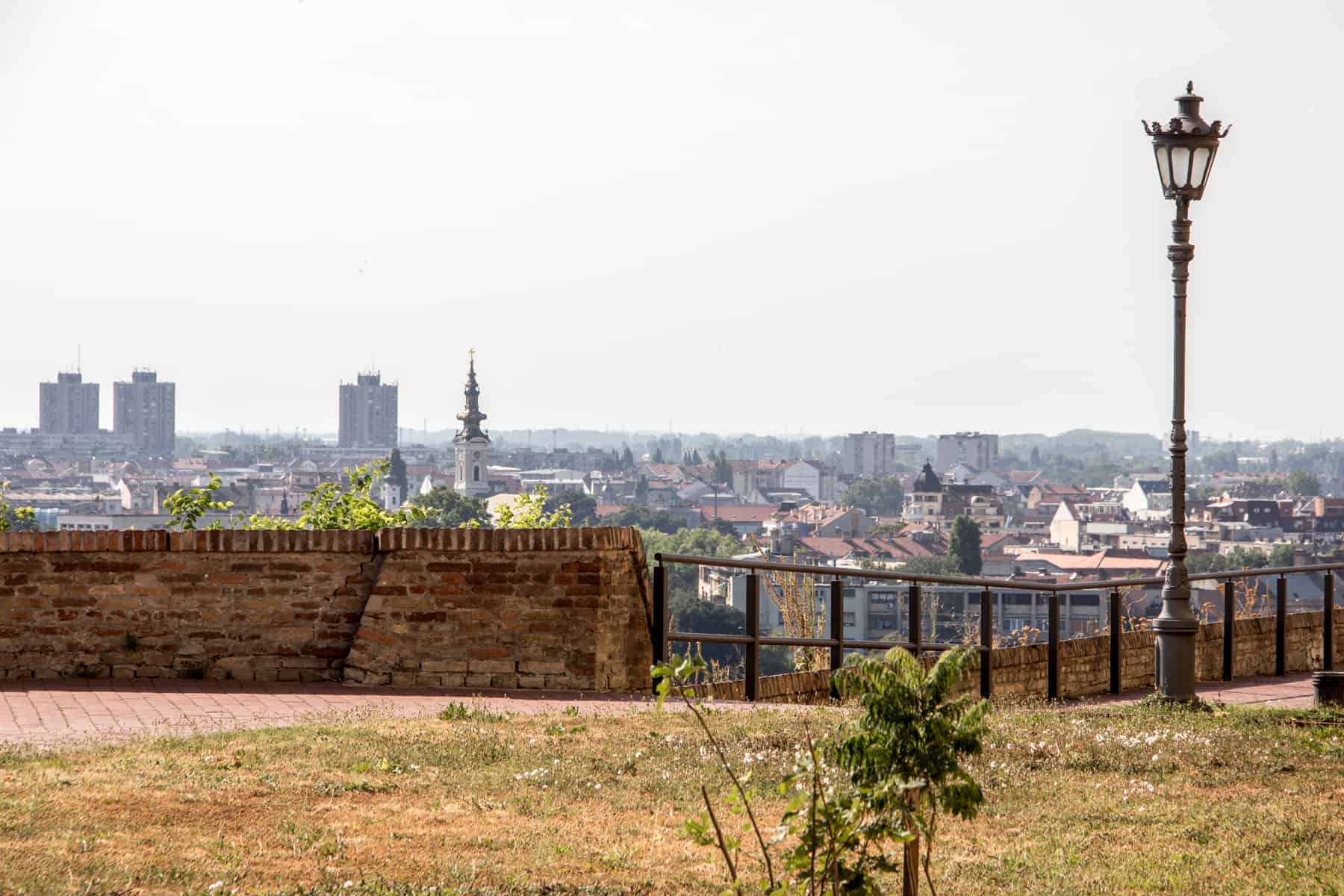 Elevated view of the Novi Sad city skyline from a grassy point with a brick wall and black lamppost in the foreground. 