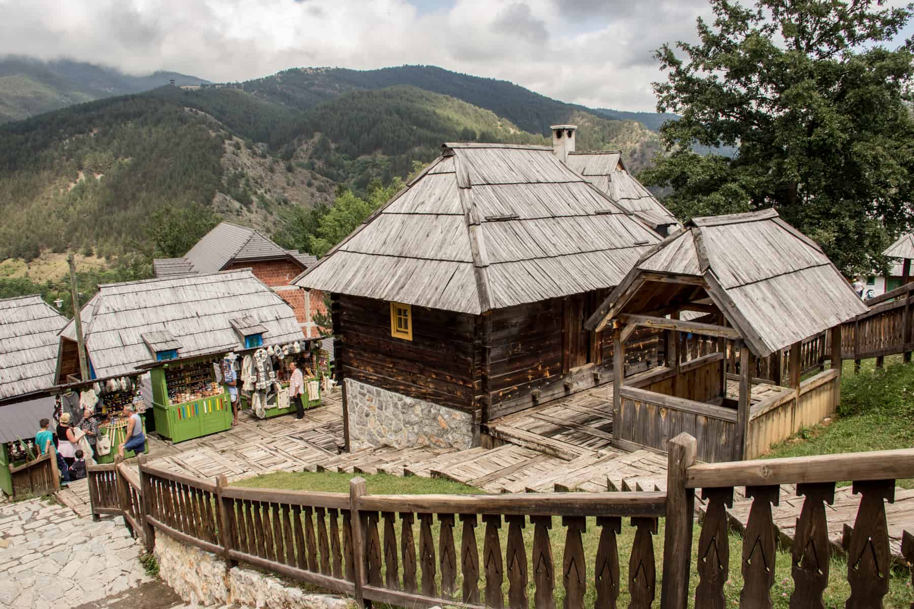 People gather around stalls at a bundle of traditional wooden houses with thatched roofs, elevated with views of the forested mountains. 