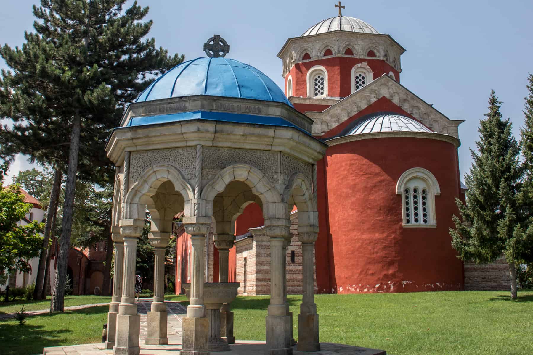 Two religious buildings stand upon vivid green grass. In the foreground, a pavilion structure with a bright blue roof. In the background, a round church painted in a bright burnt red. 