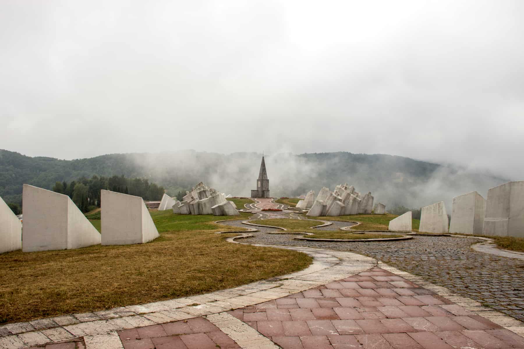 Mist hovers above a church structure hat stands on a hilltop at the end of a curling stone pathway that weaves past large and slanted white stones. 