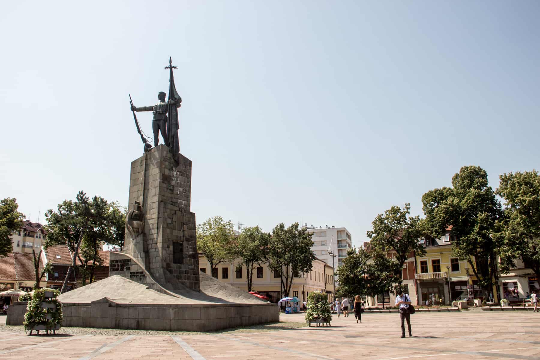 People walk through a large open square with a statue of a man holding a flag and a rifle. The square is surrounded by trees. 