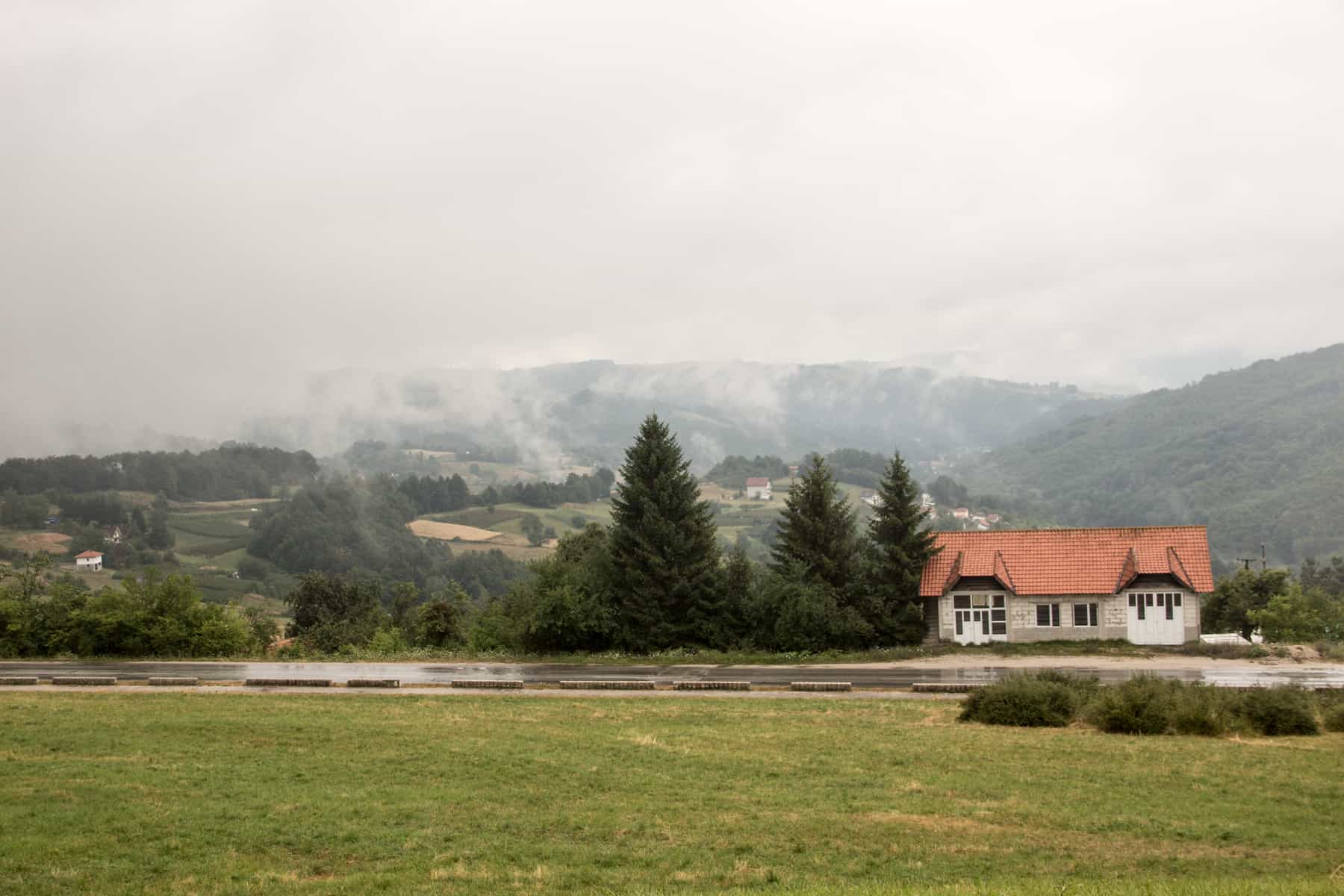 Mist rises above a green valley. In the foreground is an isolated shite house with an orange roof and a grouping of pine trees. 