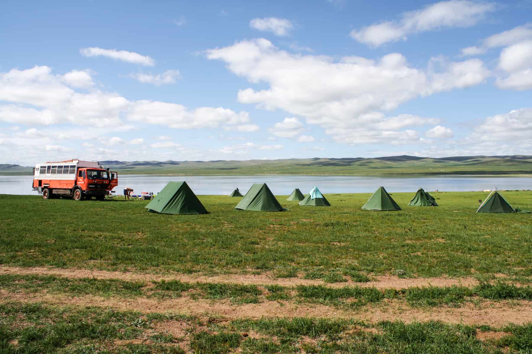 An orange overland truck and rows of green tents besides the wide Ugii Lake in Mongolia