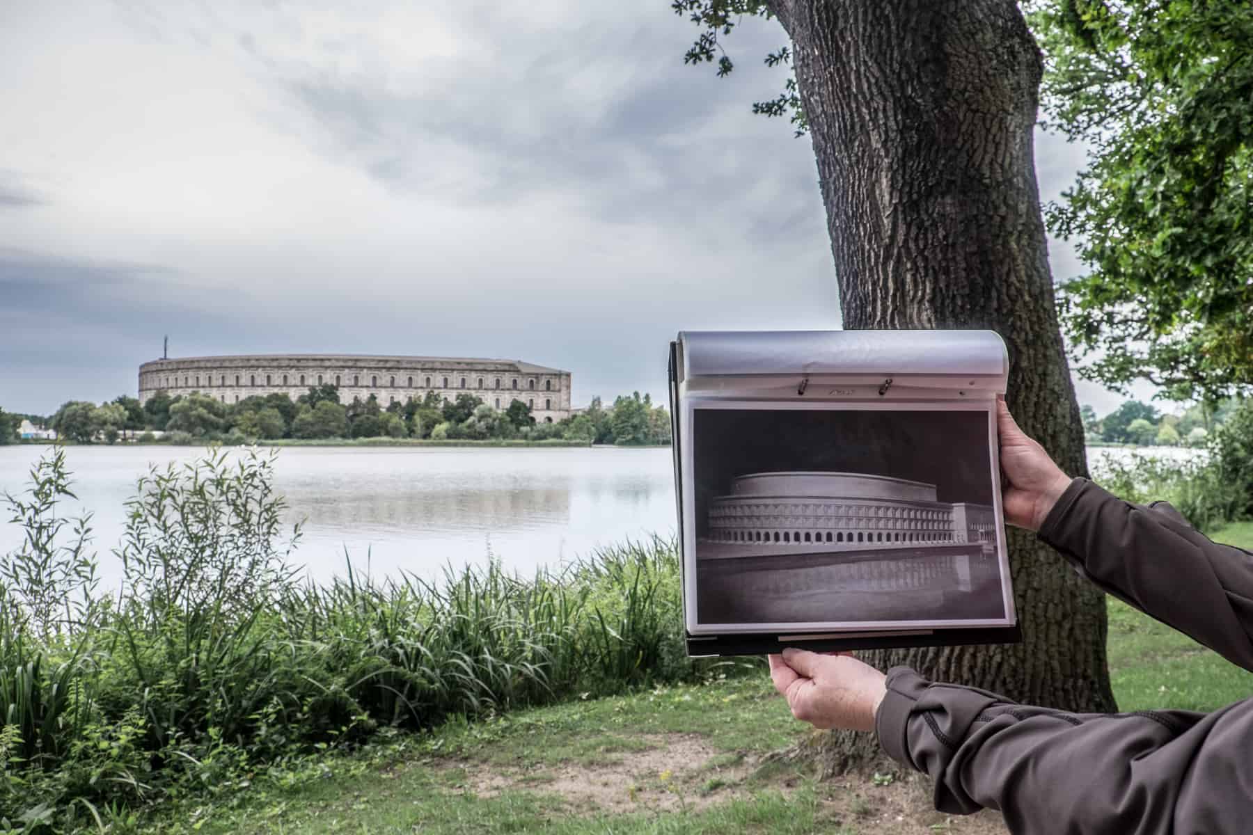 A female guide holds a picture of the finished Colosseum-like design of the Congress Hall Nazi Party Rally Ground site, in front of the unfinished construction in the background