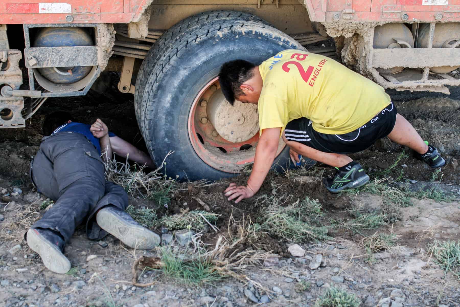 Teo men dig out a truck wheel from deep, wet mud while overlanding in Mongolia