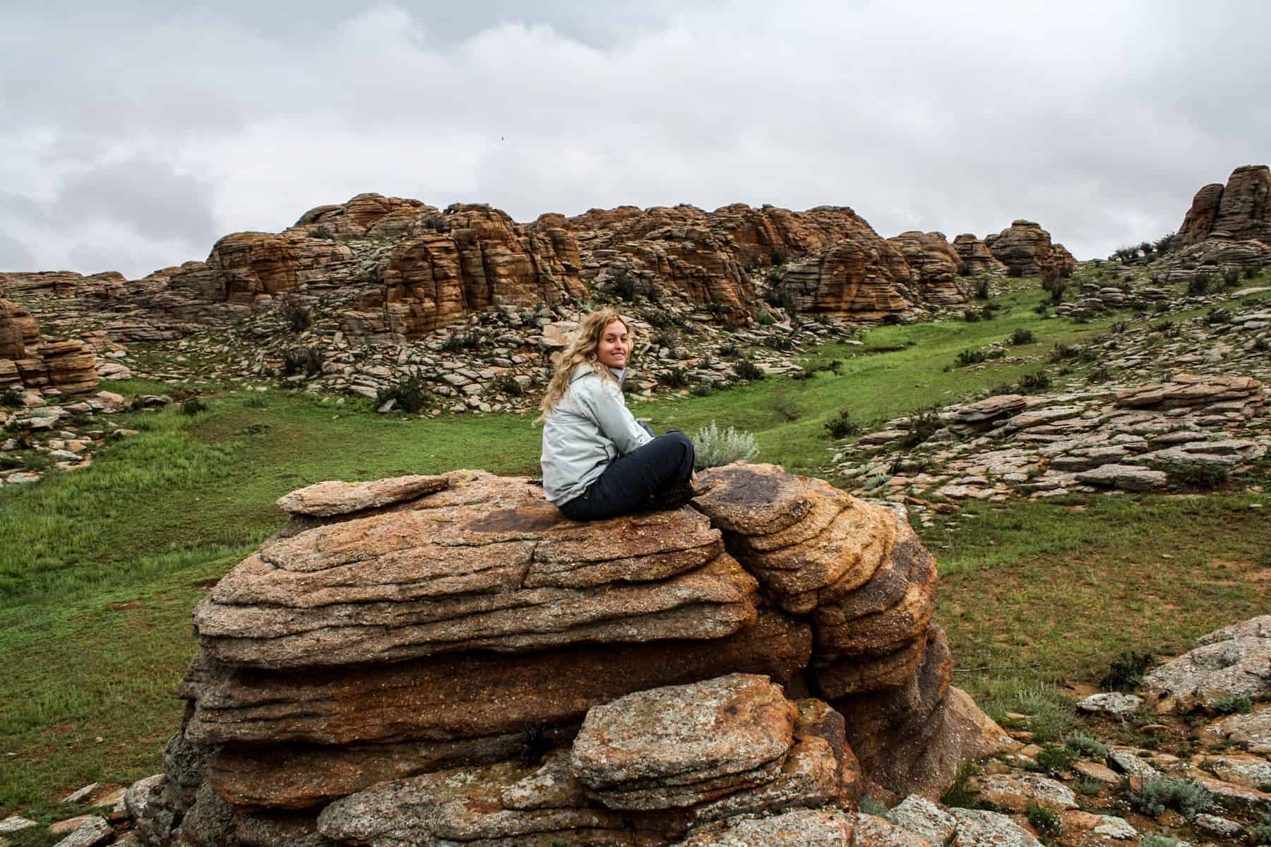 A woman sits crosslegged on top of a large rock in the middle of a grassy area, part of the Baga Gazryn Chuluu rock formations Mongolia