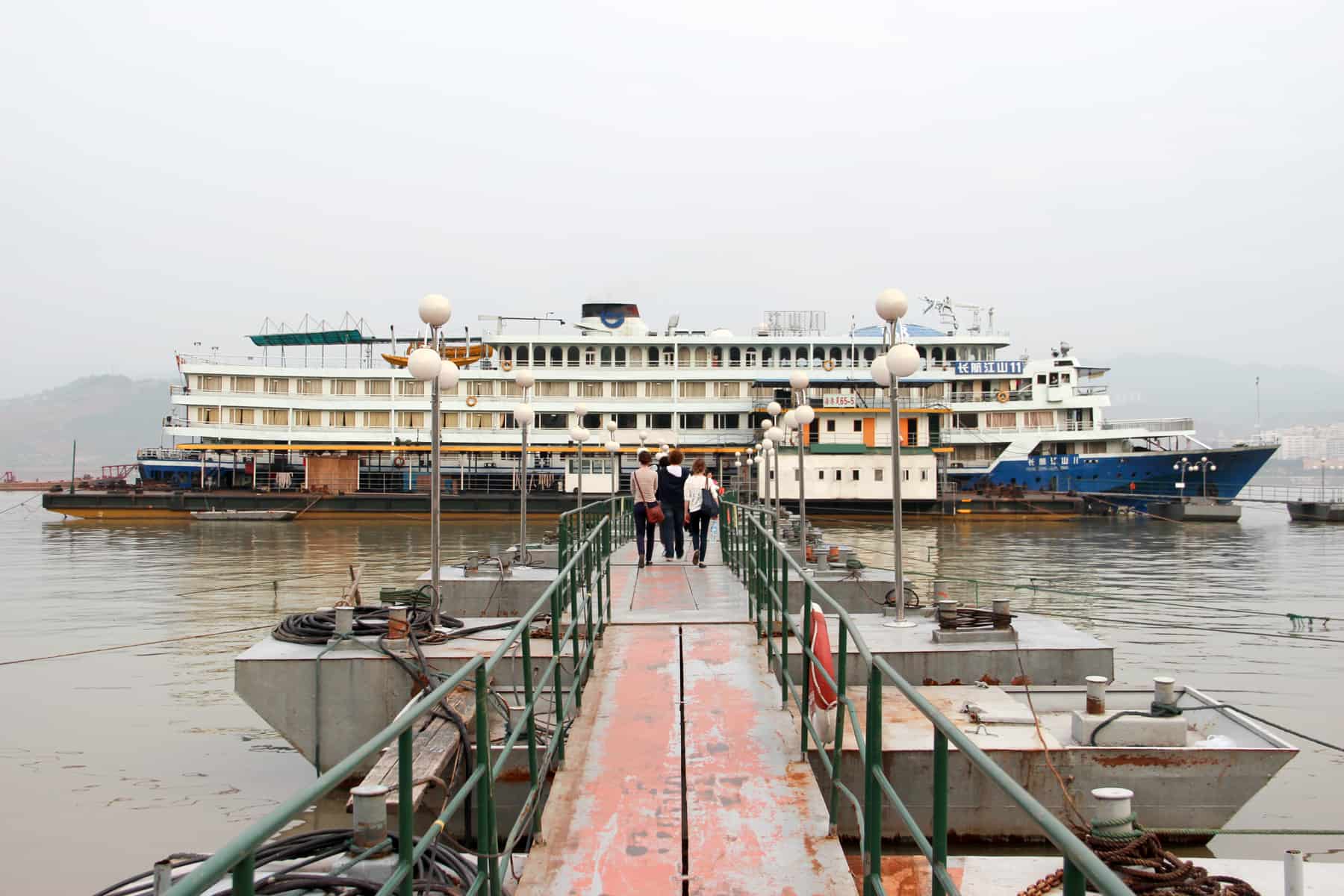 Three people walking along a worn orange pier to board a standard class Yangtze River Cruise boat