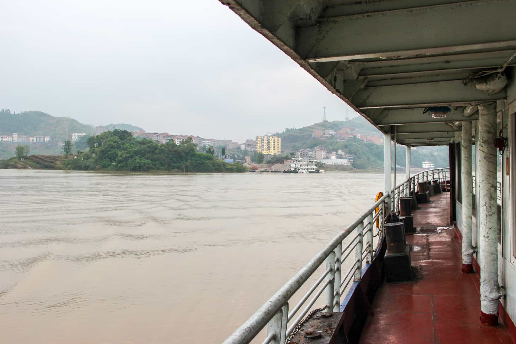 A China river cruise boat slowly floats on the dirty brown waters of the Yangtze River towards a built-up town on the hillside