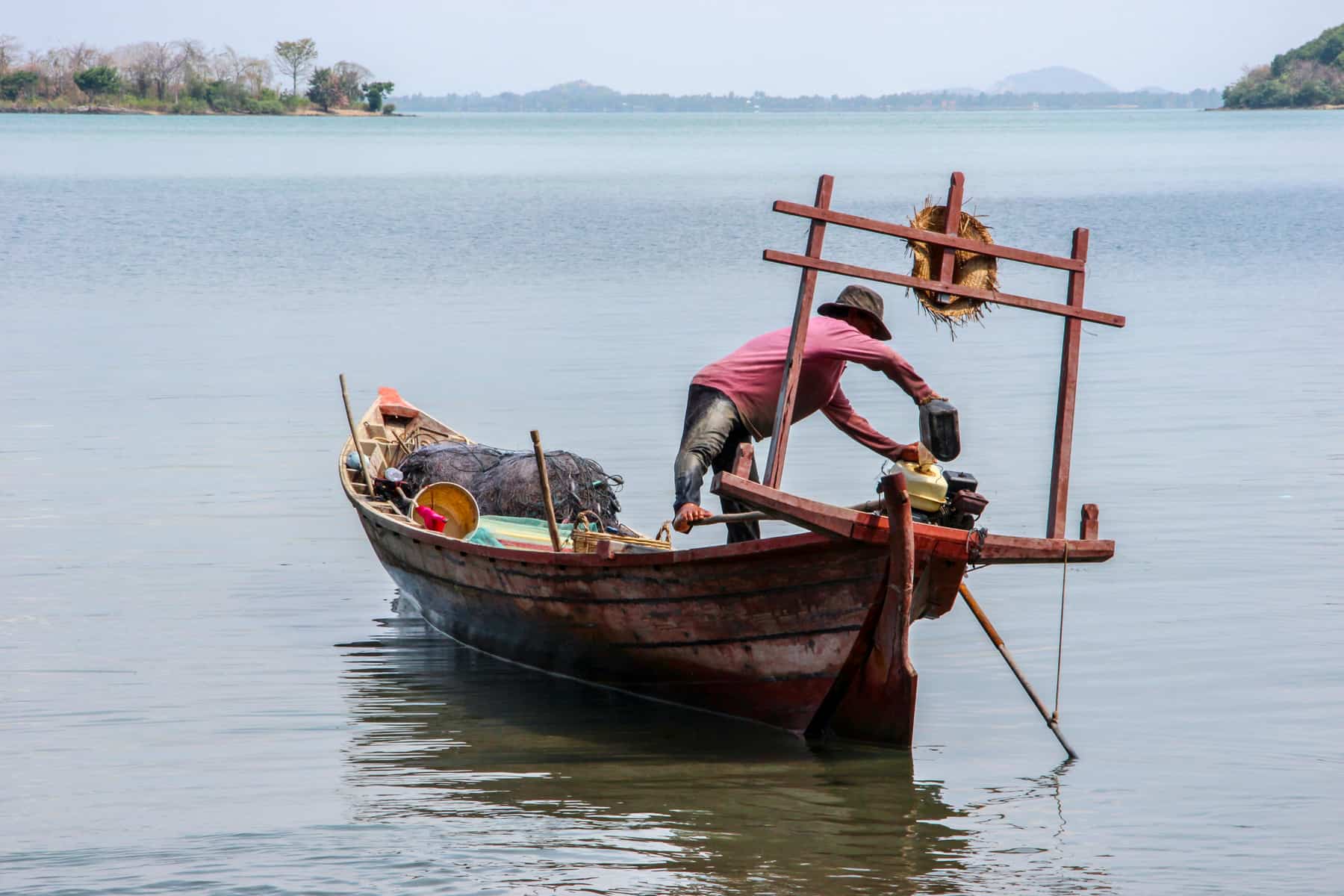 A fisherman on a wooden boat with a wooden frame, fishes while using one foot to steer the boat