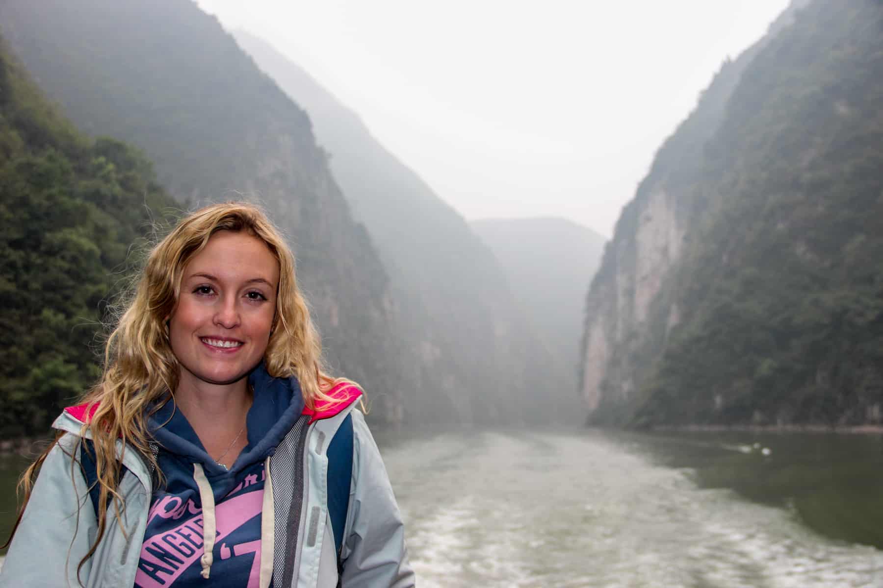 A woman in a baby blue and pink jacket stands on front of the towering, foggy gorges on a Yangtze River Cruise boat