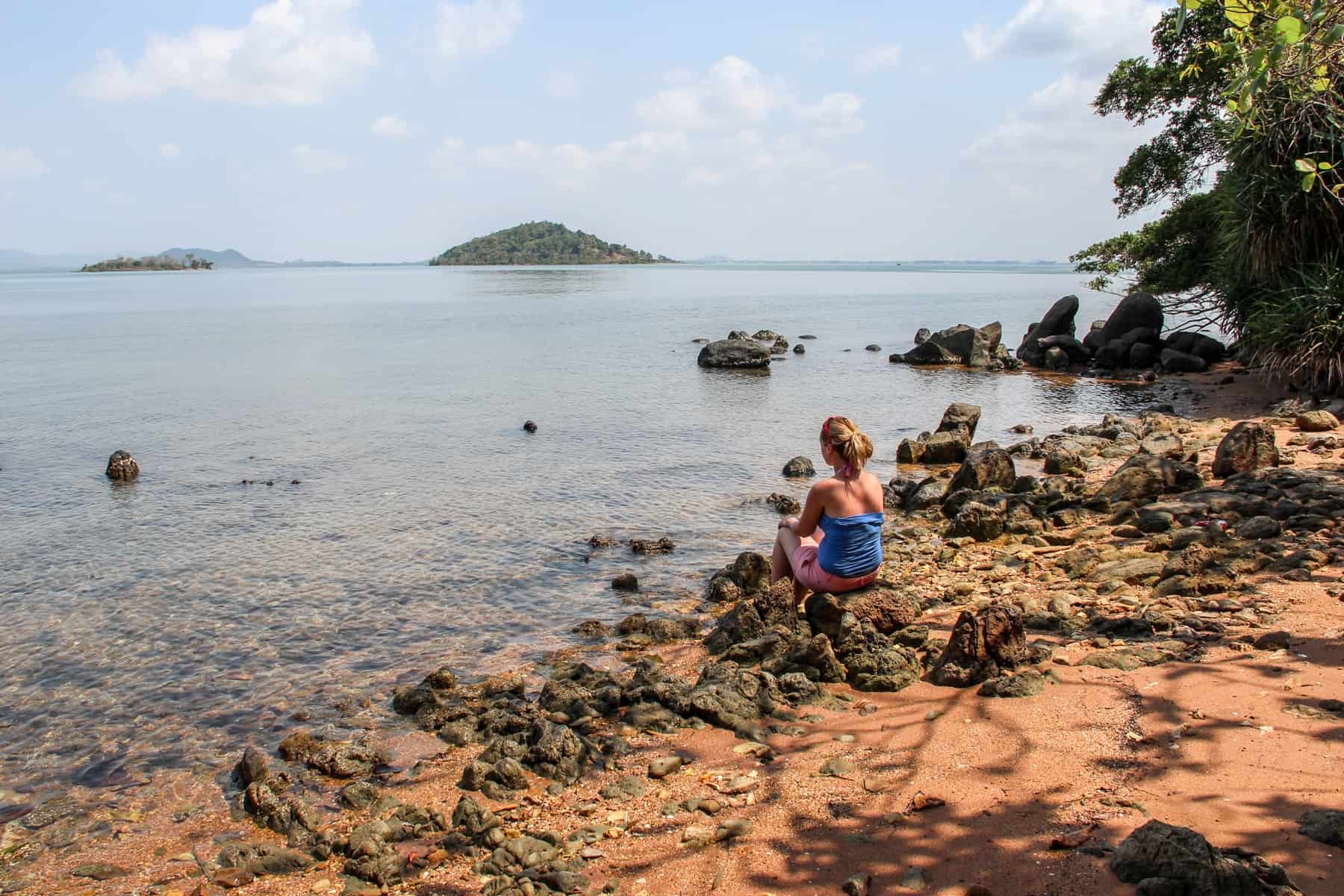 A woman sits on a rock and looks out to sea on a isolated boulder covered beach