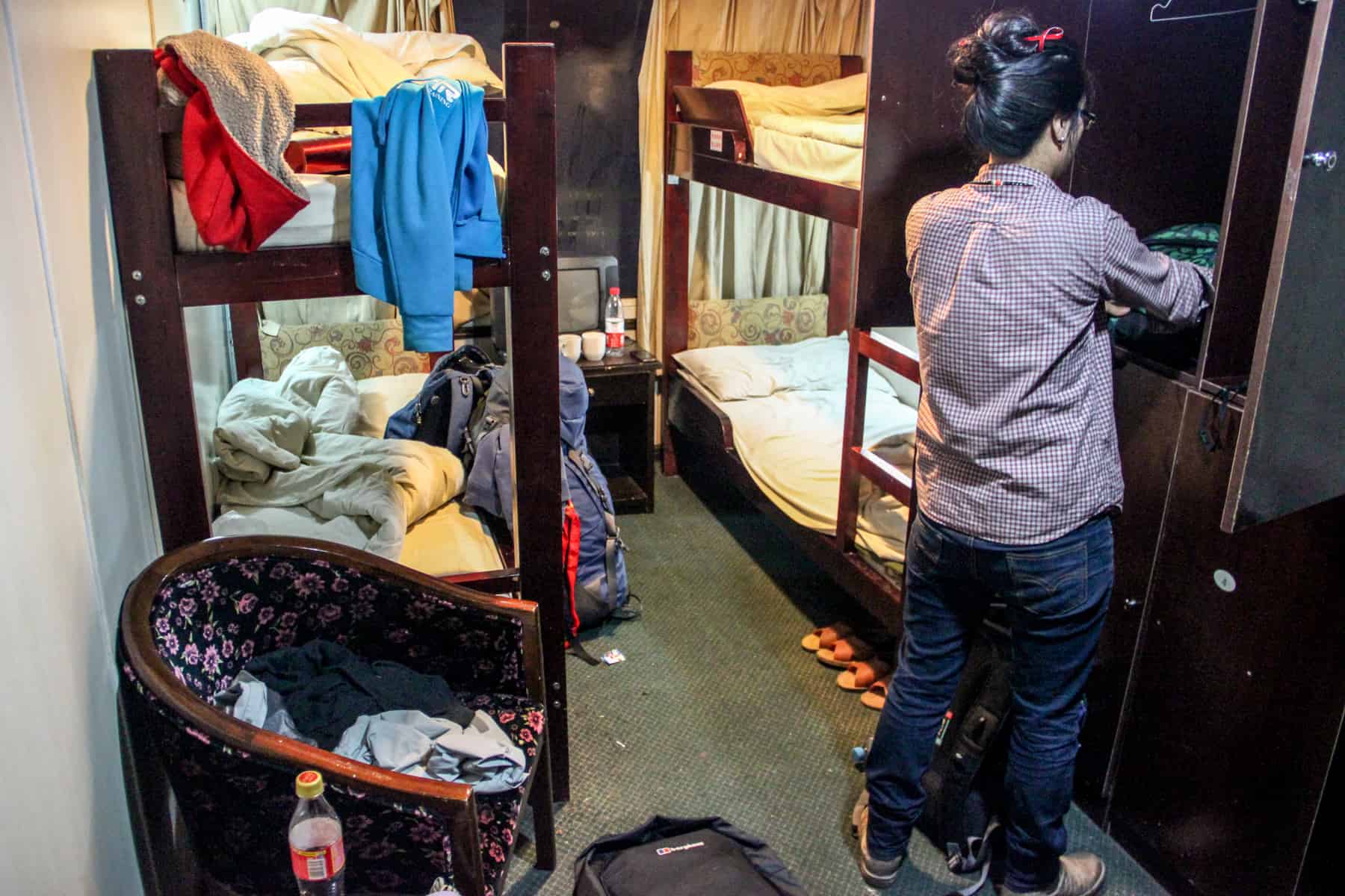 A woman folds away her clothes in a messy boat cabin bedroom with two dark wooden bunk beds