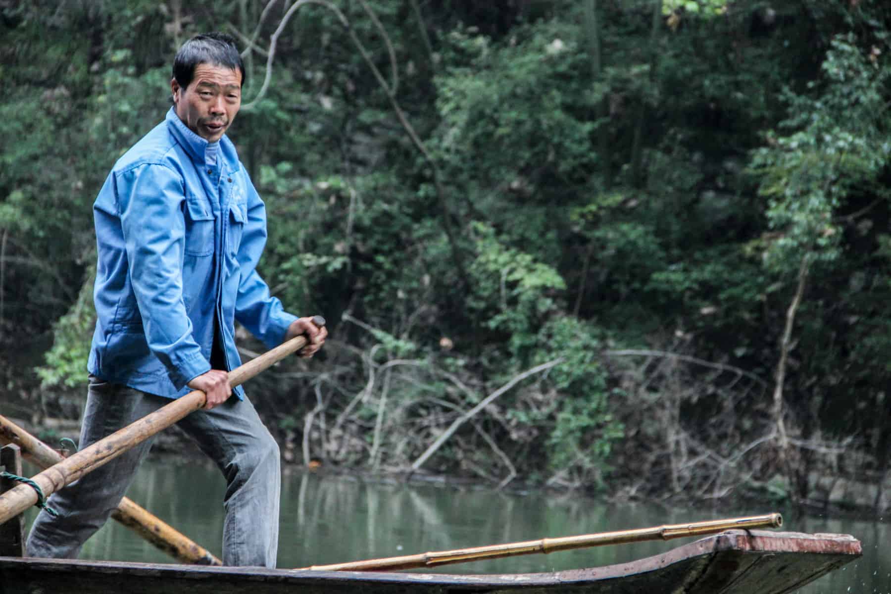 A Chinese man in a blue jacket paddles a wooden boat with a wooden oar