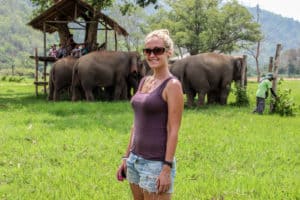 A woman wearing denim shorts and a purple vest top stands in a lush parkland in front of a herd of elephants and caretakers on a wooden platform.