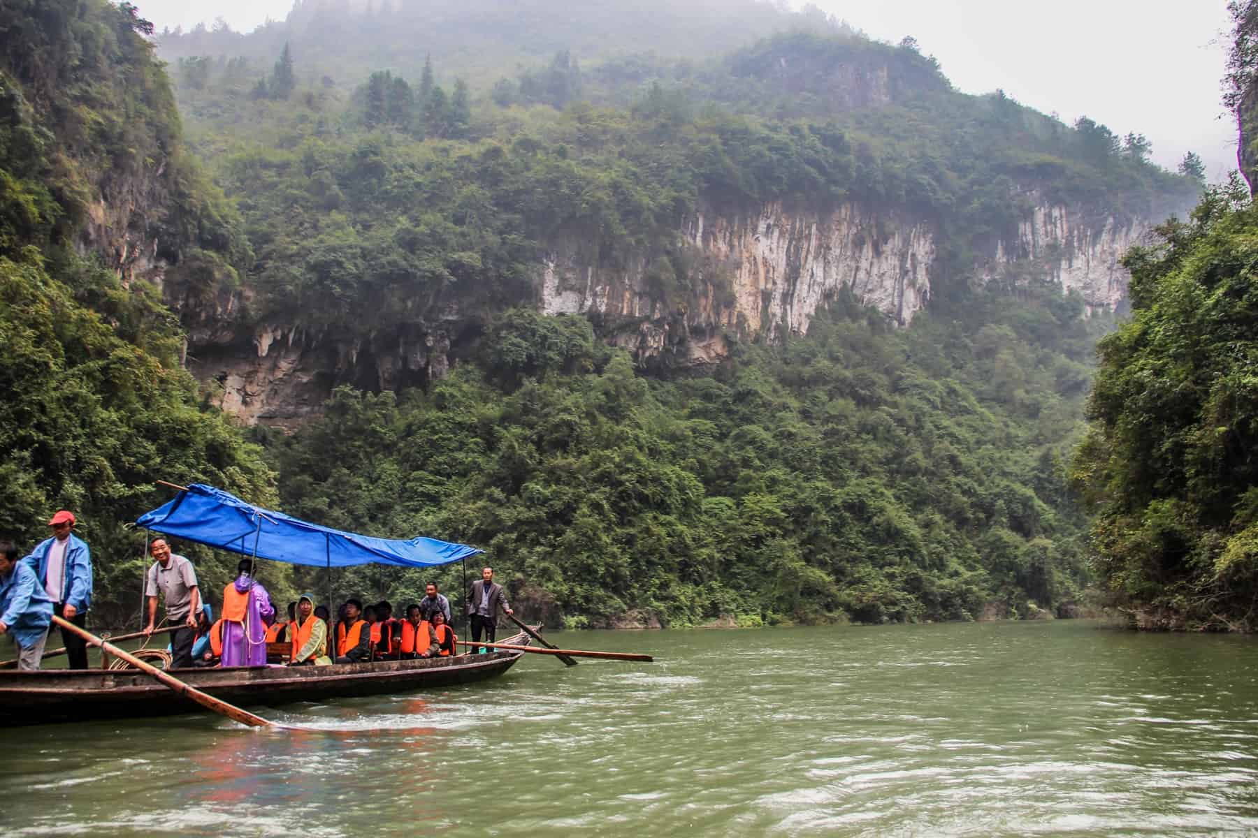 A small group of travellers in a paddle boat with a blue roof, admiring the view of the Three Gorges area on the green Yangtze River