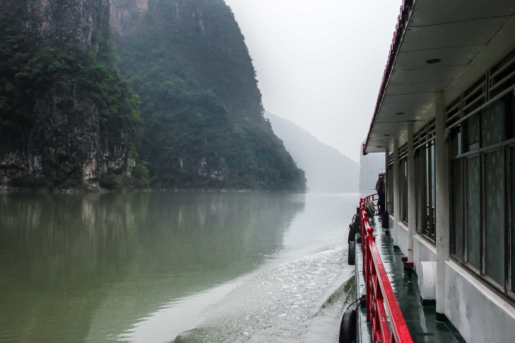 A small, white and red cruise boat slow floats on the murky green Yangtze River in China towards mist covered cliffs