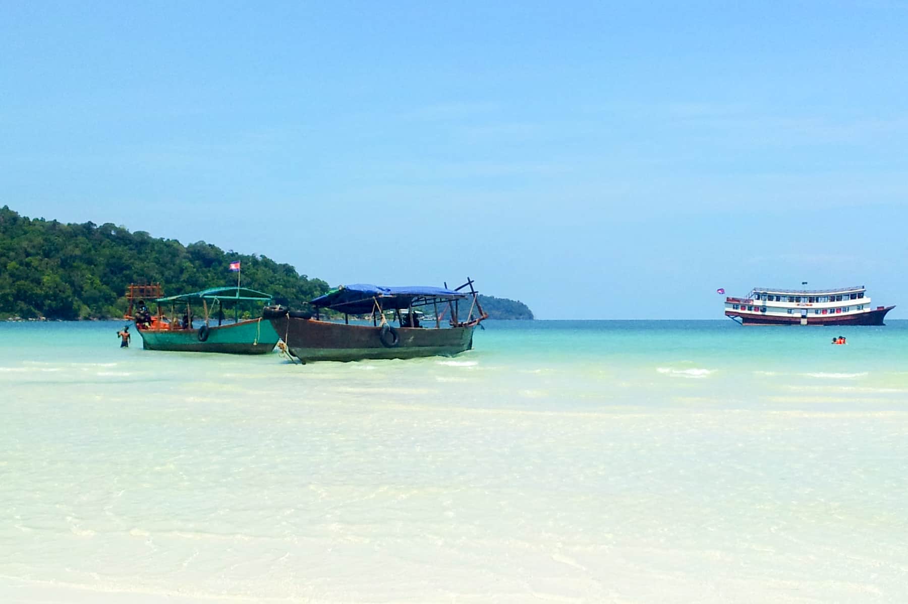 Two wooden passenger boats dock on the white sand shoreline of Koh Rong Samloem island, while another boat floats in the distance