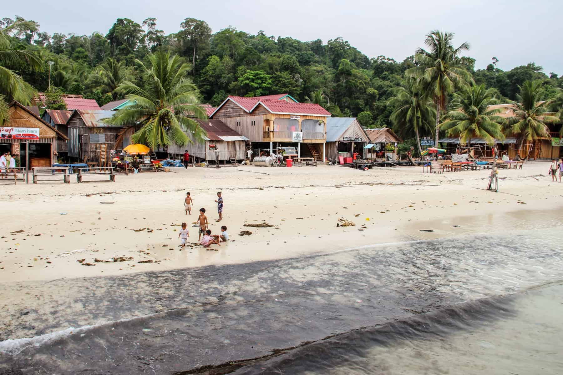 A group of six children gather on the beach of Koh Rong Cambodia, with beach huts in the background, surrounded by jungle