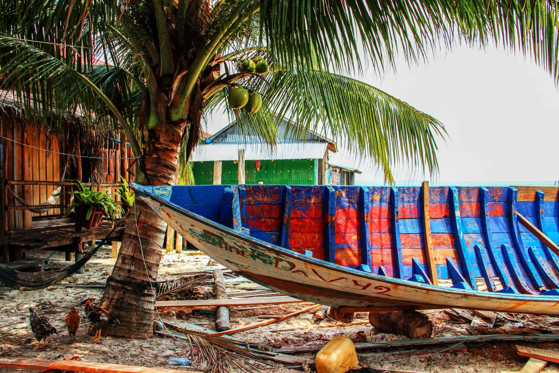 An empty blue and red painted wooden boat sits on the sand under the shade of a palm tree on Koh Rong Island