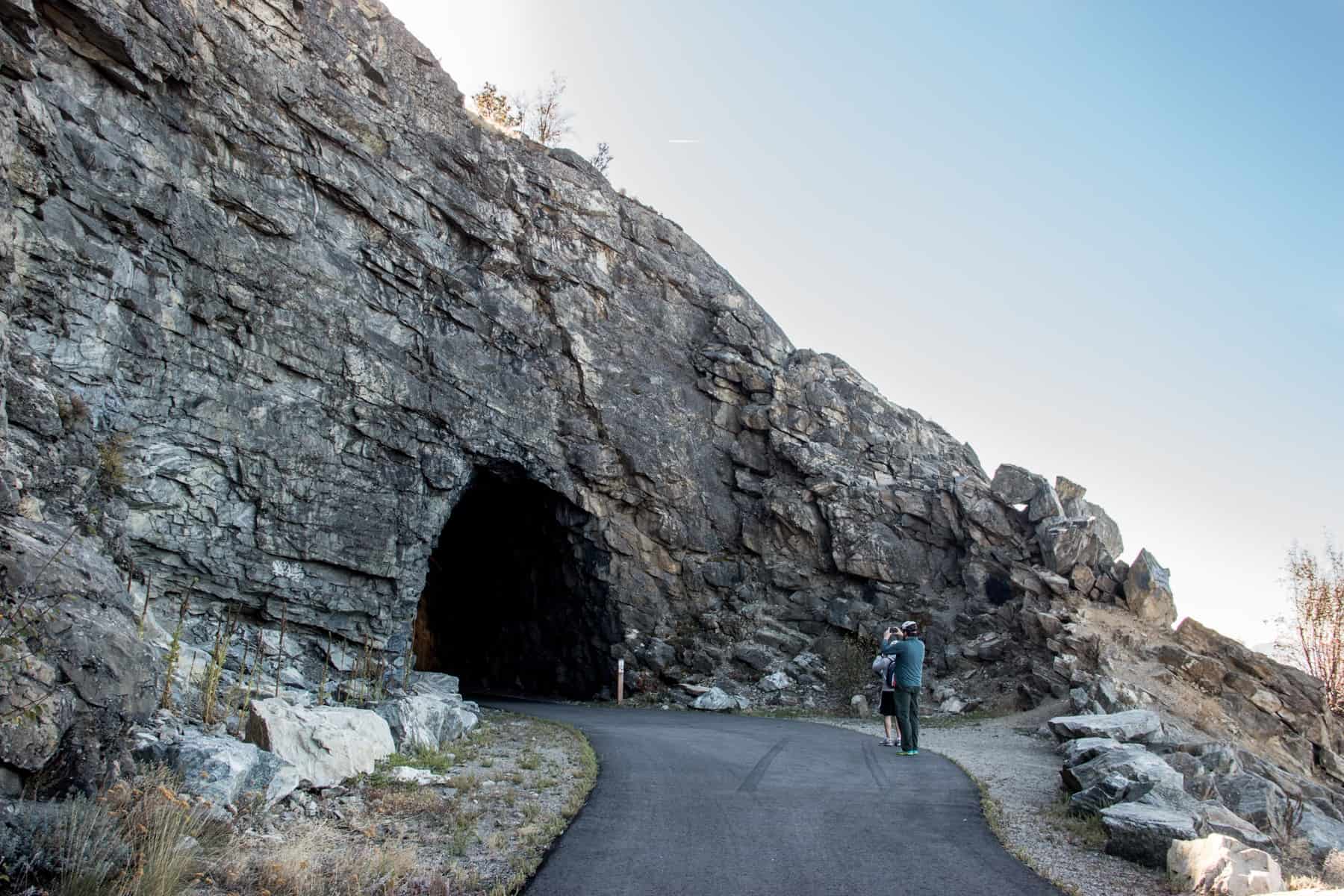 A man standing in front of a rock tunnel and taking a picture, on the Myra Canyon Trailhead on the Kettle Valley Rail Trail.