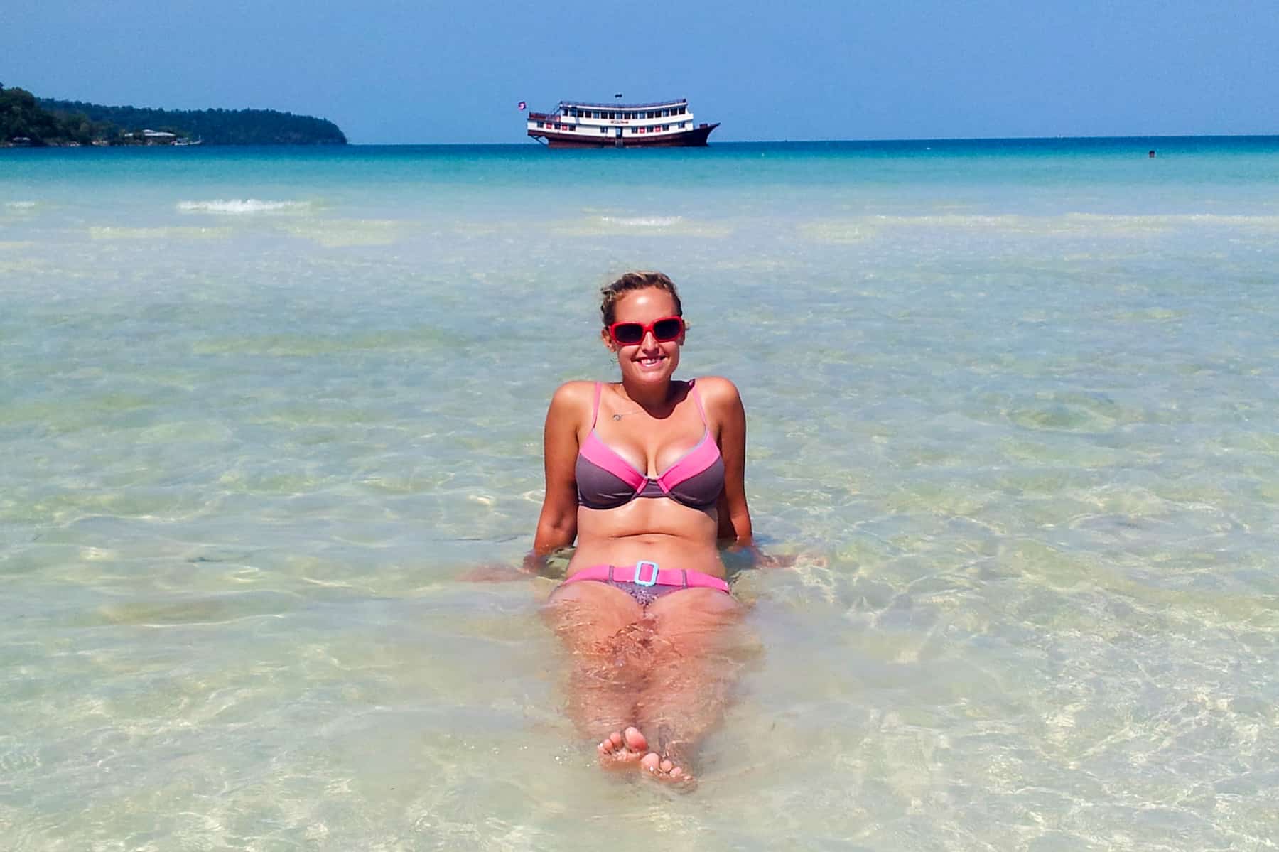 A woman in a bikini sits alone in crystal clear waters on the beaches of Koh Rong Samloem in Cambodia. It's sunny and a passenger boat flaots away fro the island in the distance