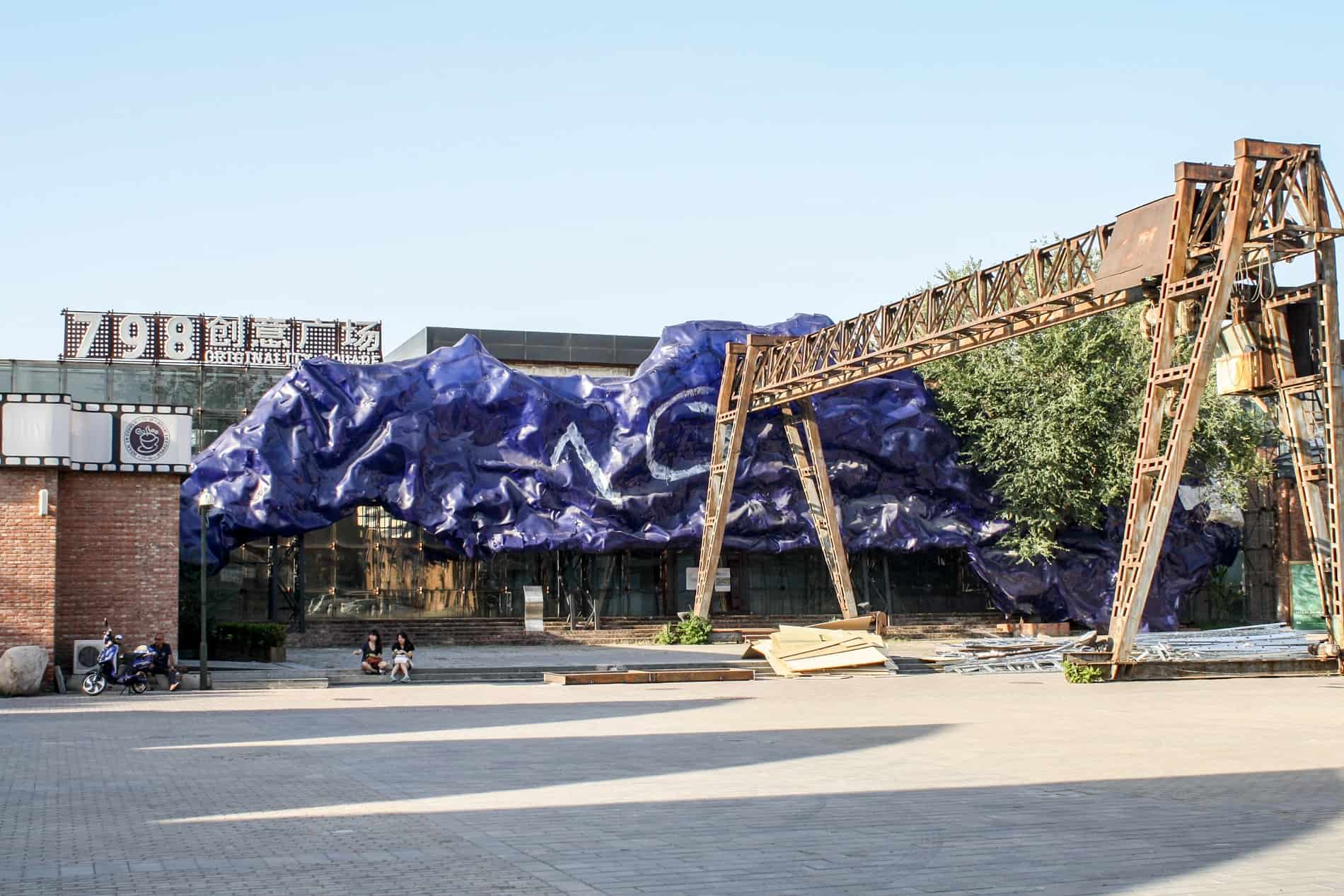 Two people sit outside The 798 Art Zone in Beijing marked by a purple cloud sculpture and steel framework. 