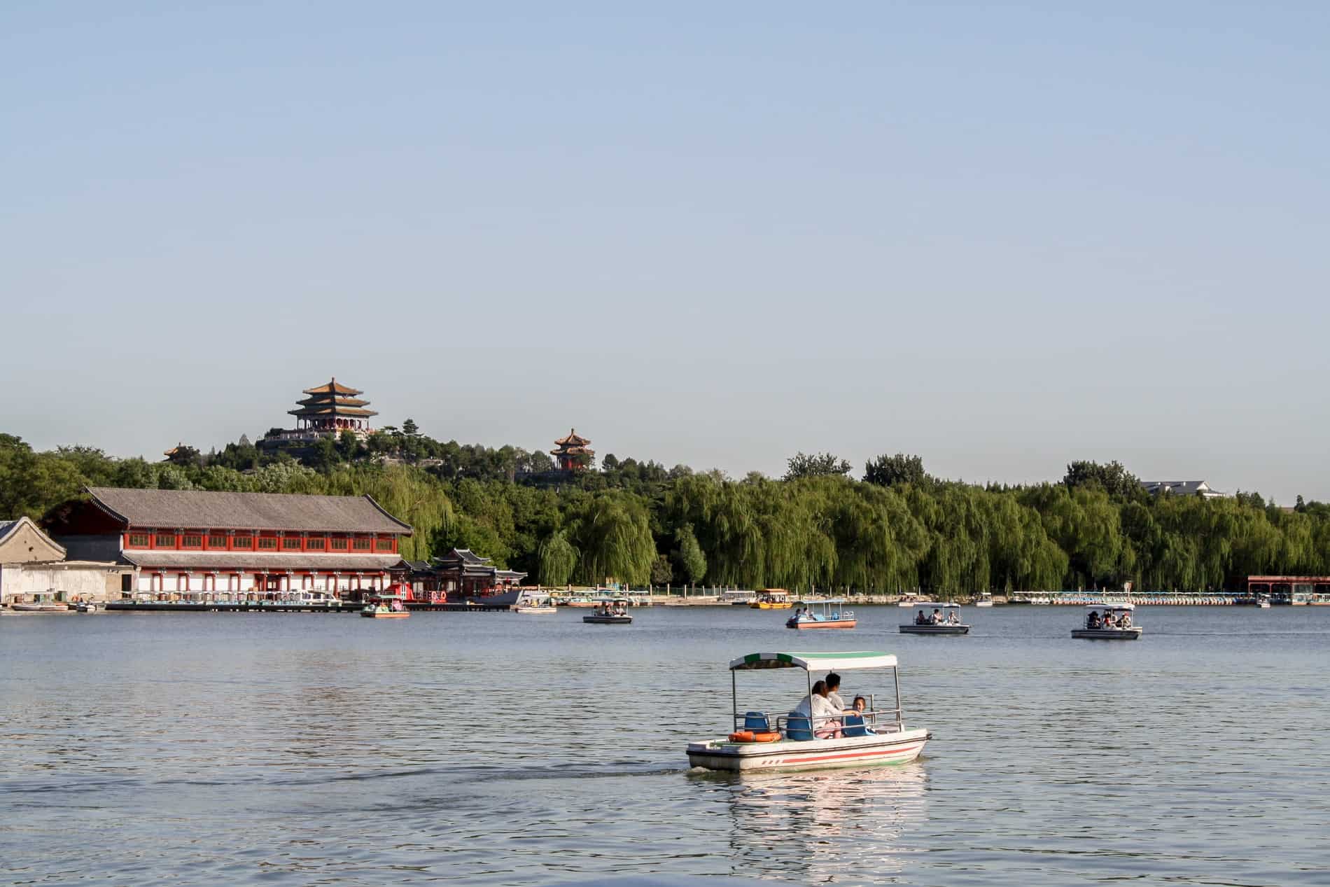Boats on a lake in Beijing next to a park dotted with tall tiered pagodas. 
