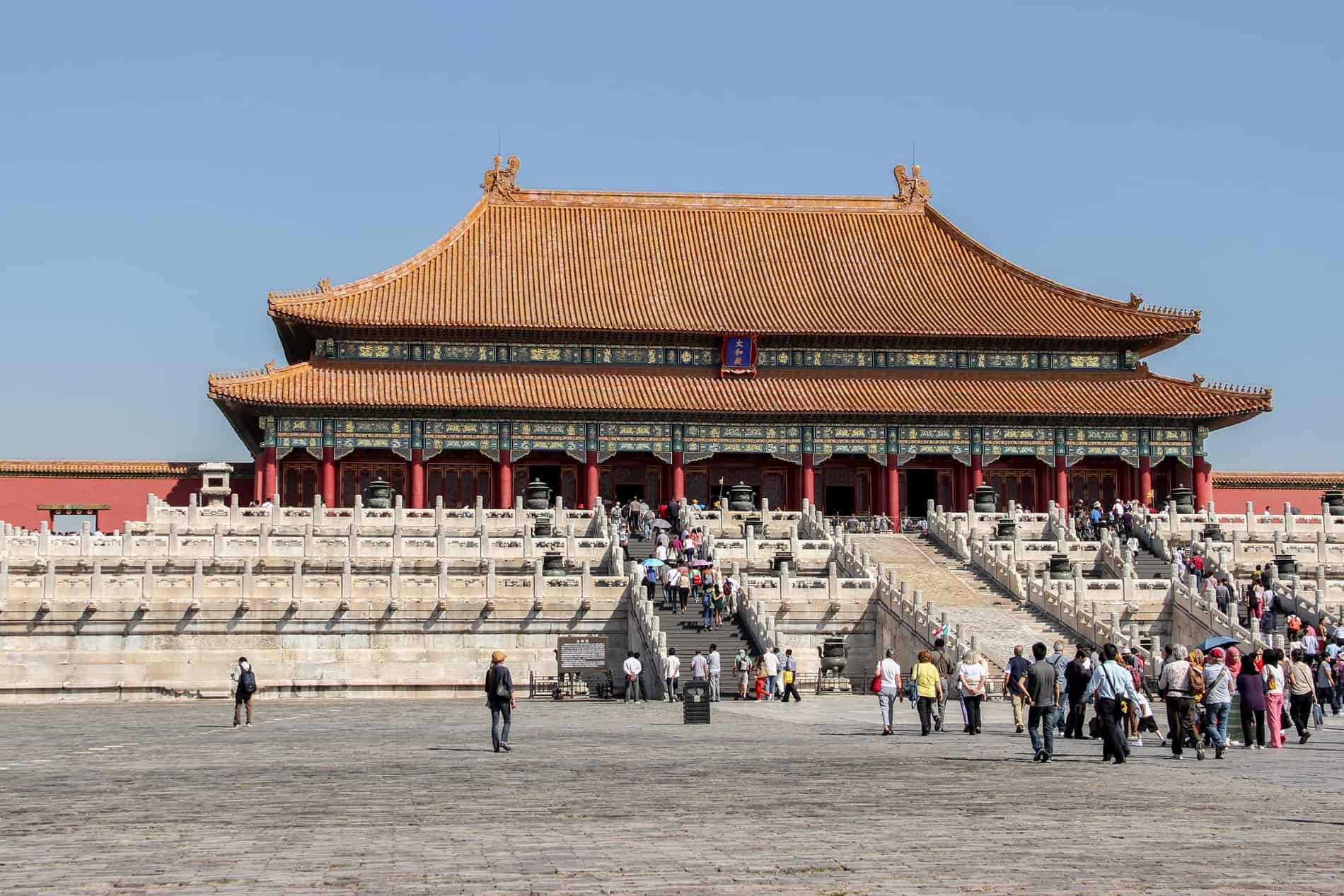 Groups of people walk towards and up the stone steps of a Chinese pagoda style building in the Forbidden City palace complex in China. 