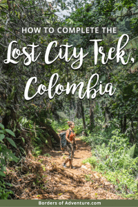 A woman in an ornage top and green trekking pants stands on an ornage mud pathway in the middle of the thick green jungle on the Lost City Trek in Colombia