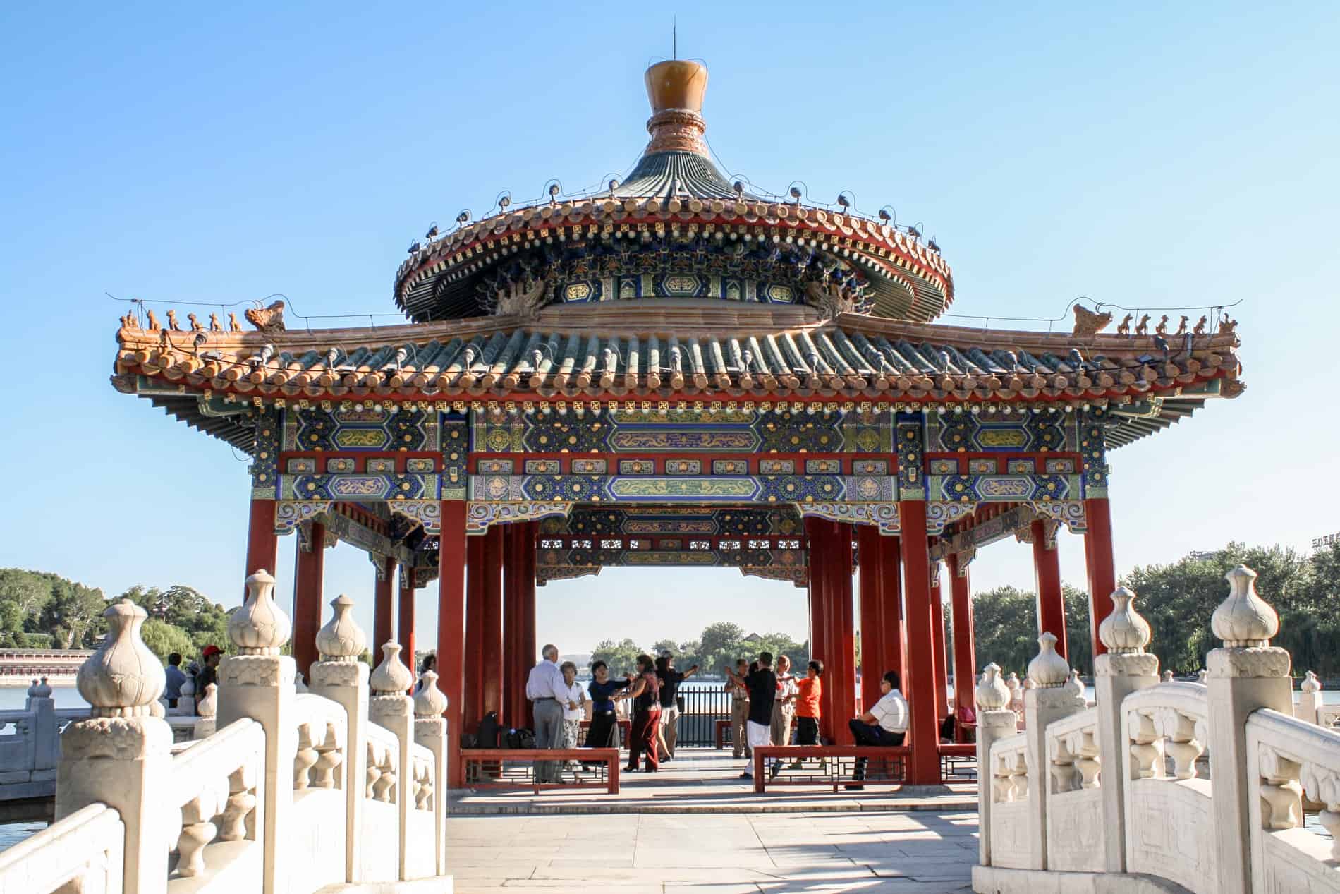 People standing underneath a traditional chinese pavilion with multicoloured paintwork, next to a lake. 
