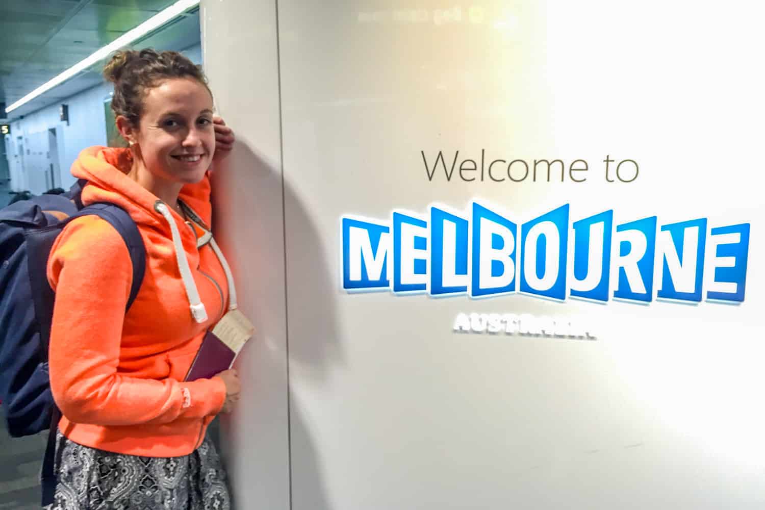 A woman in a bright orange top and black and white pants stands next to an airport arrival sign that says "Welcome to Melbourne Australia".