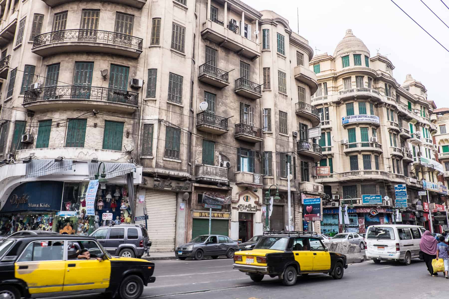 Yellow and black taxis pass by the tall light coloured buildings on a busy city street in the Mediterranean style city of Alexandria in Egypt