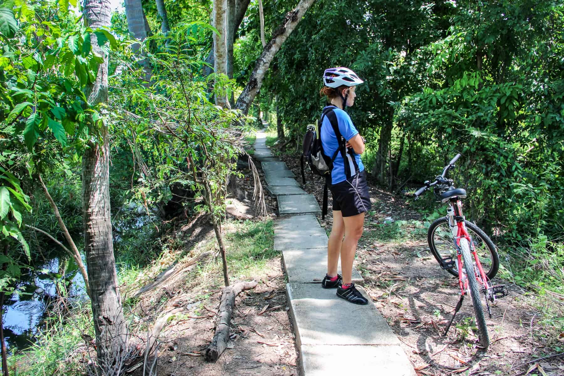 A woman in a blue t-shirt and black shorts takes a break from biking in the Bangkok countryside, taking shade on the pathway surrounding by green bushes
