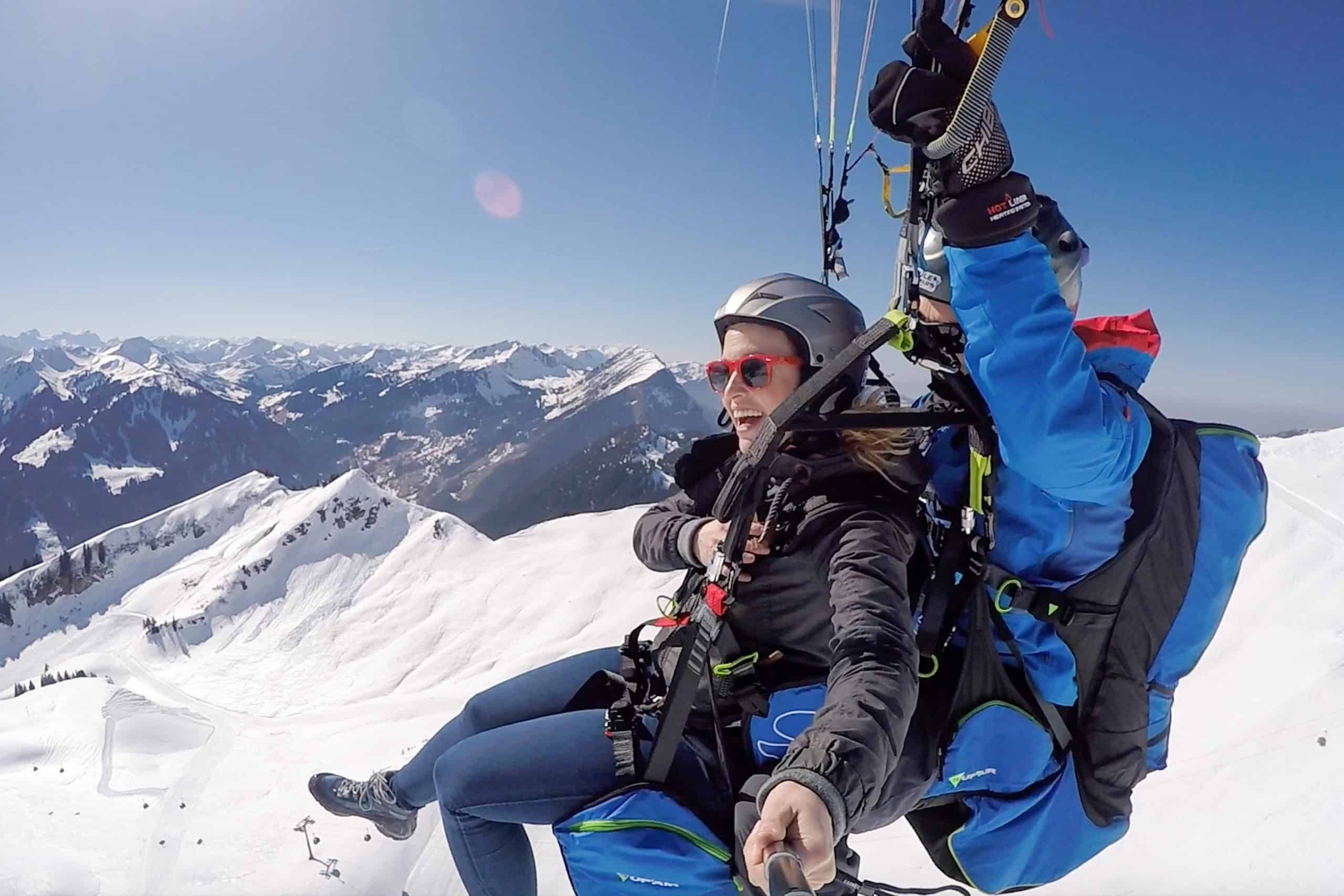 A woman paragliding in Austria with a male pilot behind her and the snow peaked Austrian Alps in the background