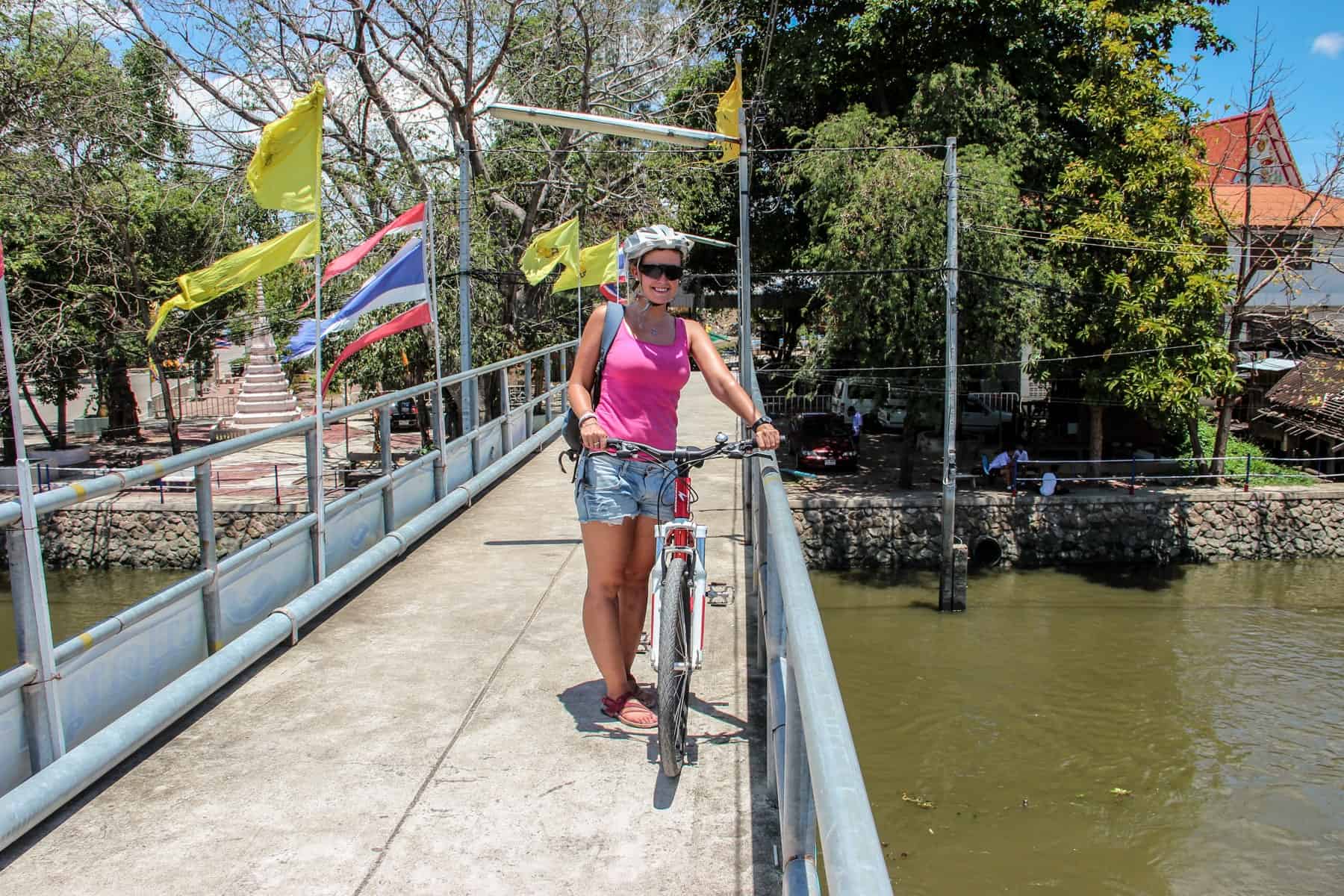 A woman in a pink top and demin shorts stands with her bike on a concreate bridge next to a small river in Bangkok