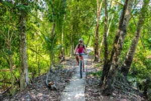 A woman wearing a pink top and demin shorts riding a bike on a narrow stone path through a thick green forest track on a bike tour in Bangkok