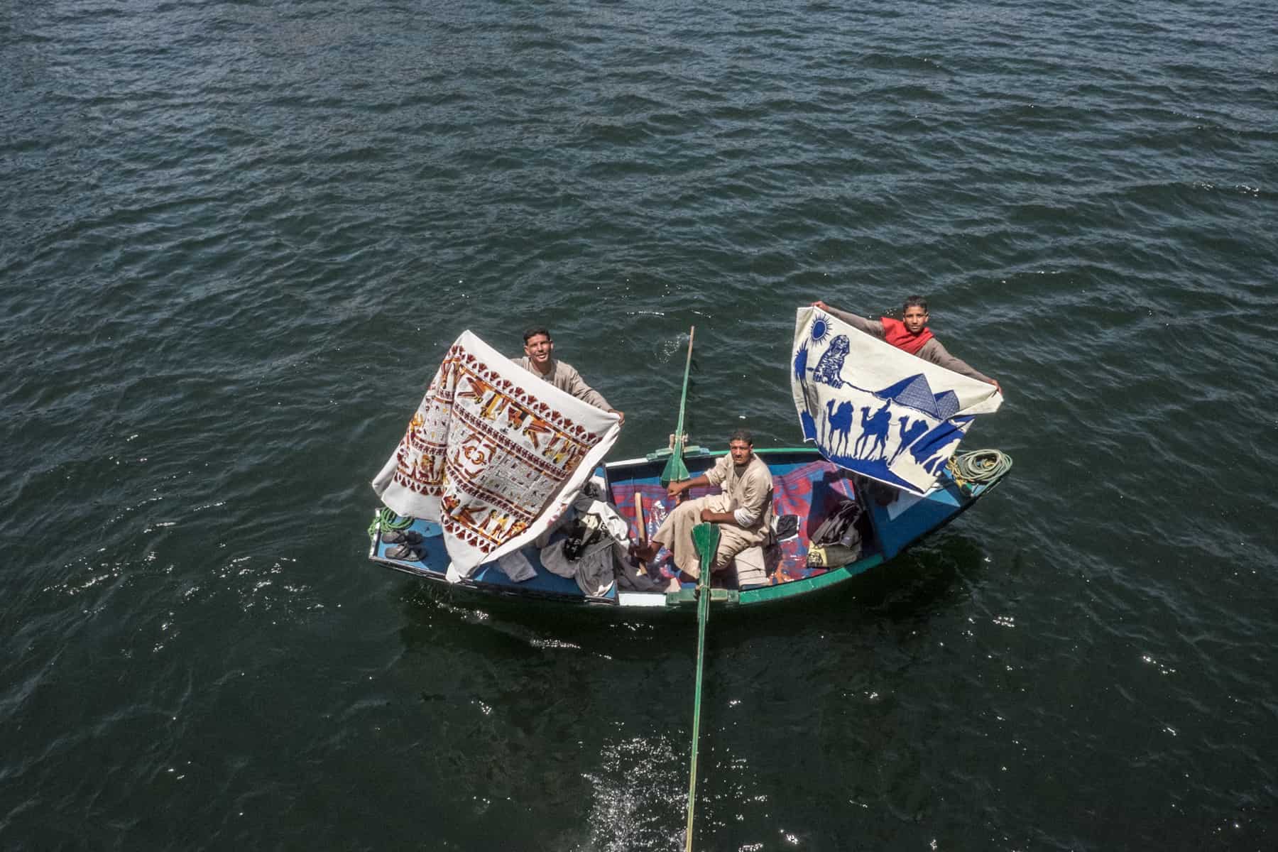 Three men in a wooden boat on the River Nile in Egypt. Two hold up patterned material trying to sell to passengers at the boat window