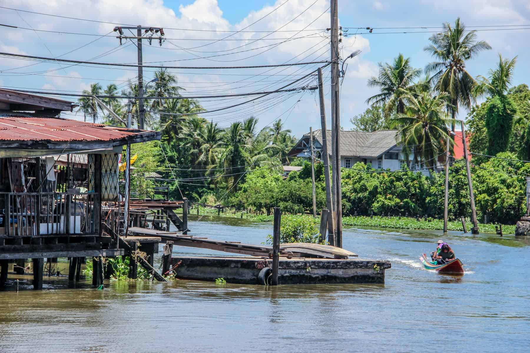 A wide river estuary in rural Bangkok with a floating wooden stilted house on the left and green jungle on the right