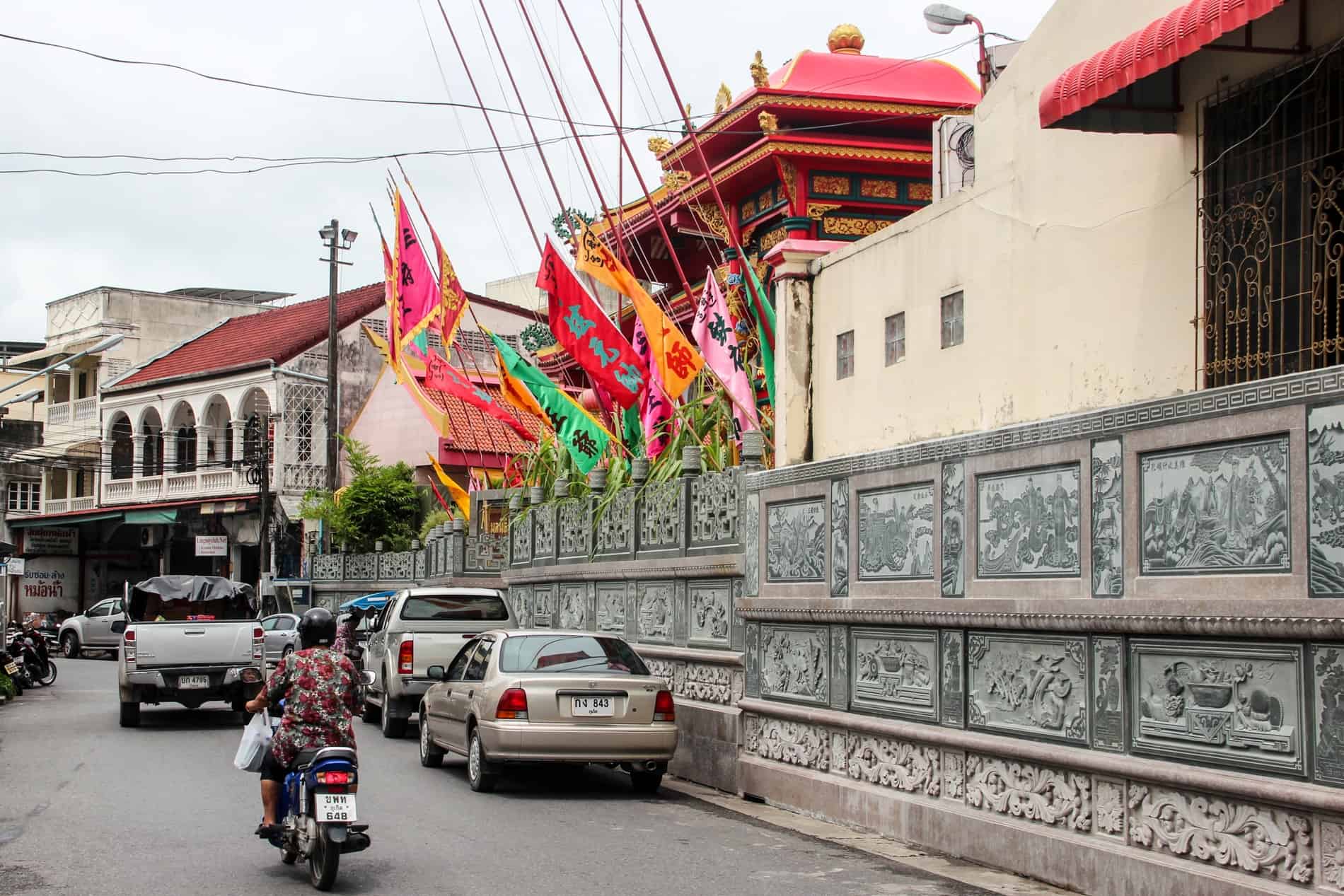 A woman on a motorbike and parked traffic on a street in Phuket with a red-roofed Chinese temple. 