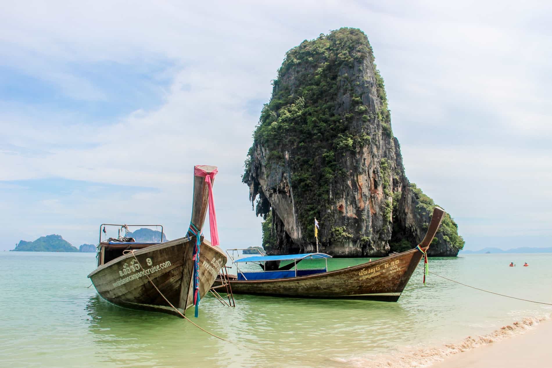 Longboats in Thailand moored on a beach in front of a limestone formation. 