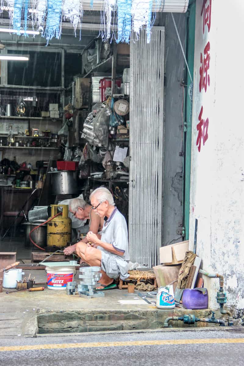 Two elderly men work on their craft in an open workshop space in the old town of Kuching, Sarawak