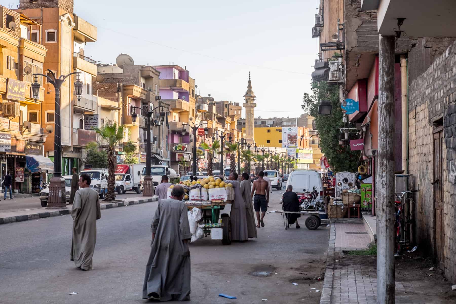 Egyptian men wearing long grey tunics walk past a fruit stall vendor on a wide golden lit apartment lined street in Luxor, Egypt