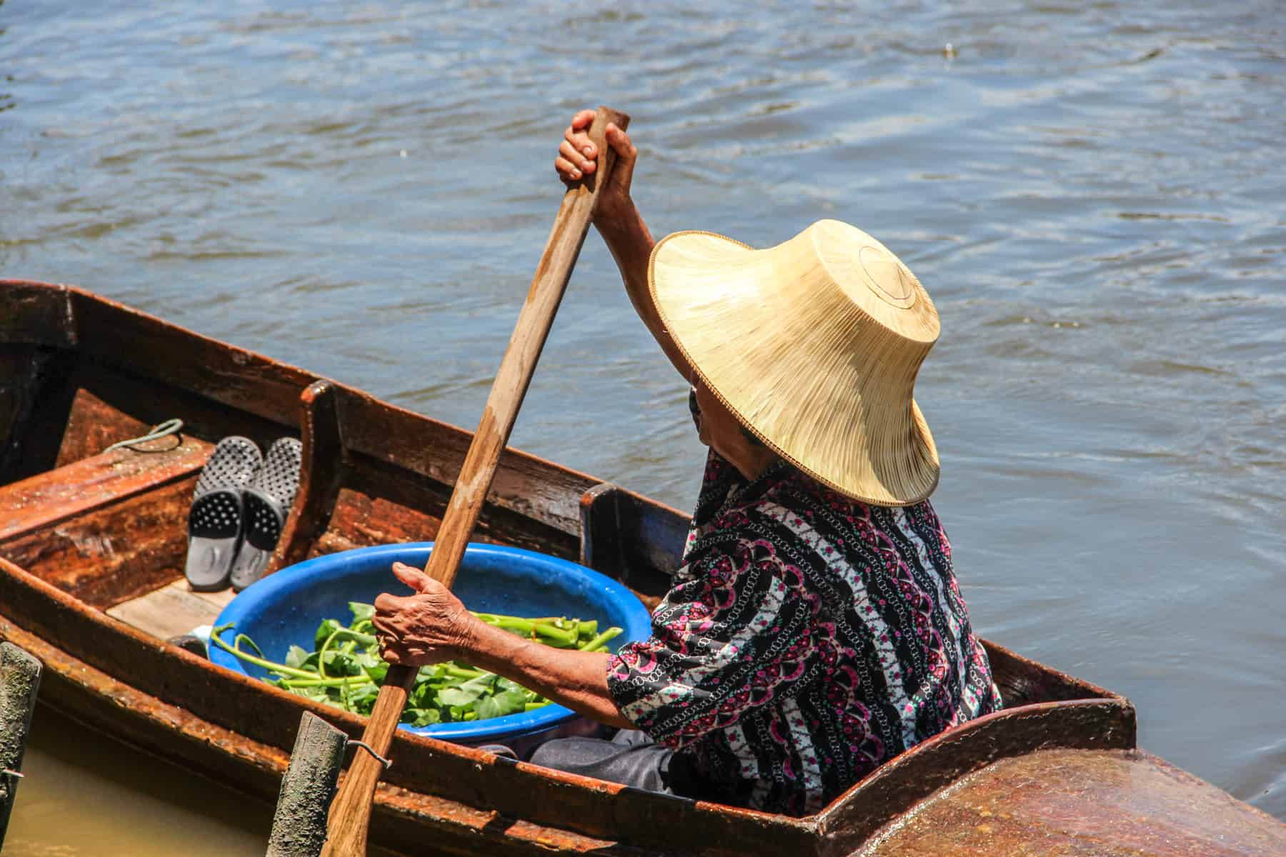 A Thai lady in a straw sun hat sells green vegetables from her wooden boat on the river in Bangkok