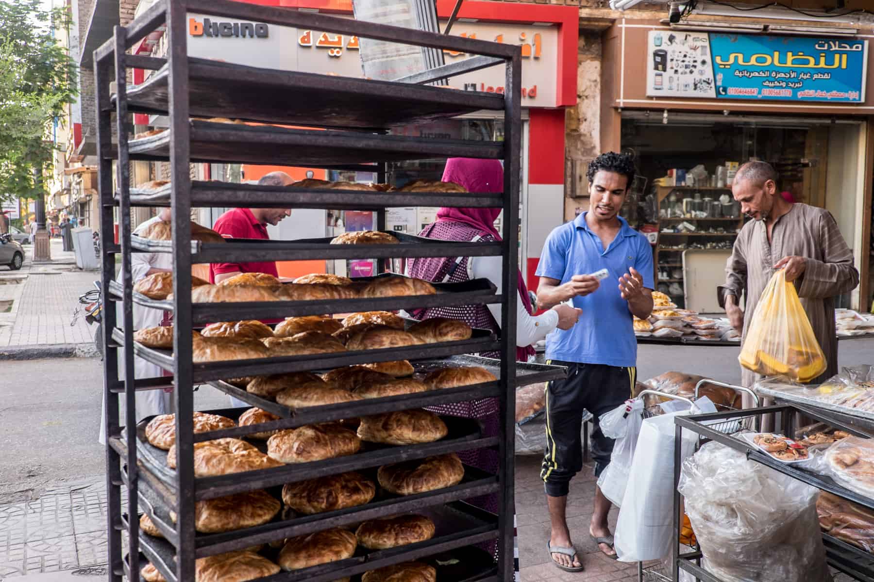 A man in a blue t-shirt exchanging money with a customer, standing next to a tall shelving rack stacked with freshly baked bread.