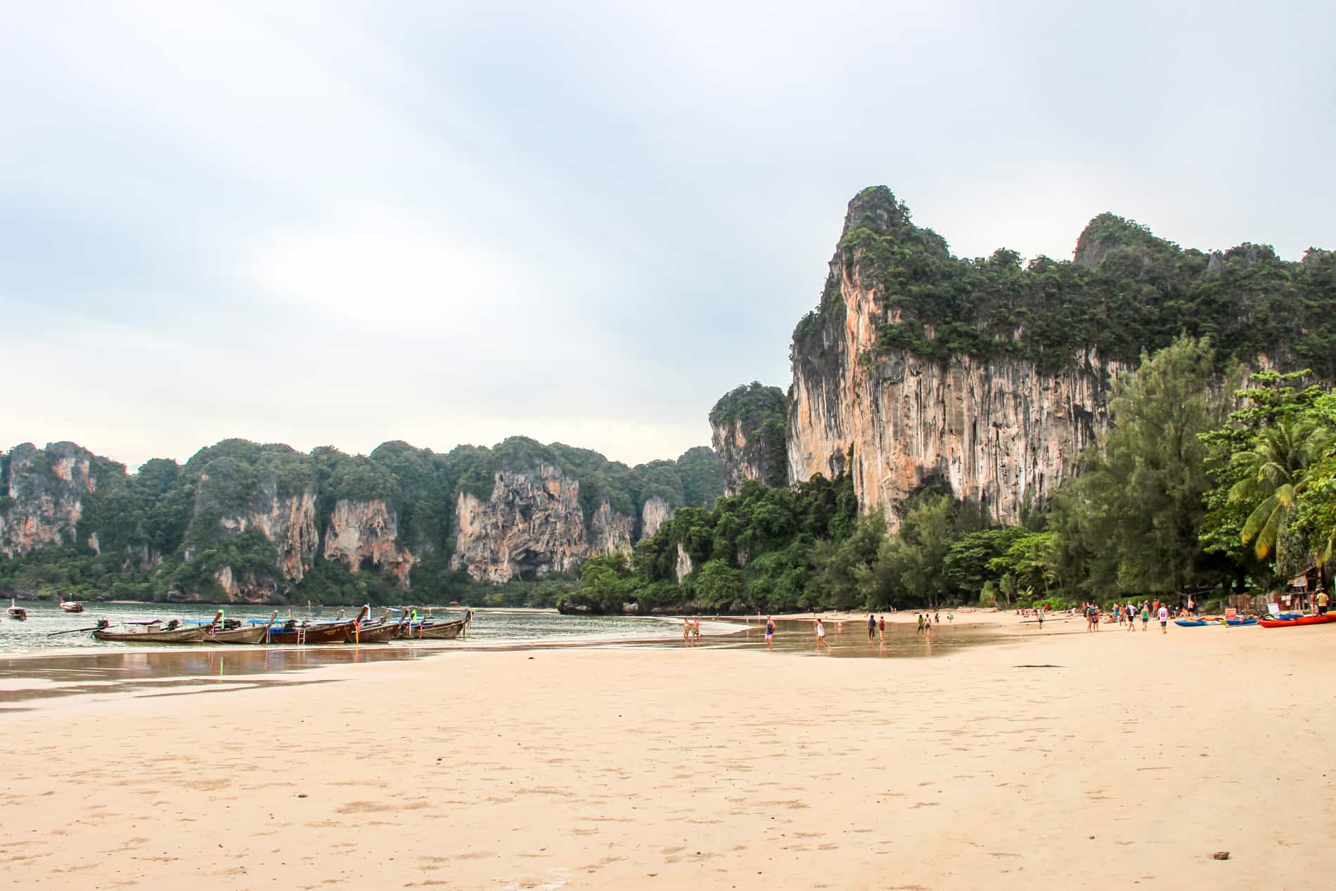 Wooden long boats and people on a limestone cliff-backed beach in Krabi, Thailand. 